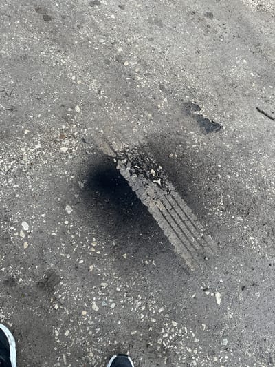 In Galiche, Bulgaria, an overhead view captures a section of weathered asphalt or concrete pavement. The primary subject is a distinct black tire mark, approximately 35-40 centimeters in length, showing multiple parallel tread lines. A concentrated black smudge, indicative of rubber transfer or heavy friction, is present at one end of the mark. The grey pavement surface contains scattered light-colored aggregate and minor debris. A dark shadow partially obscures the upper left portion of the tire mark. In the lower frame, the toes of a person's black and white athletic shoes are visible, indicating an individual standing over the scene. No other individuals or significant objects are present.