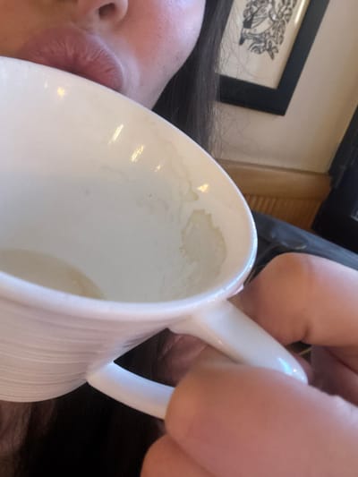 A white ceramic mug with a handle is held close to the face of a person with dark hair and fair skin. Residue lines the inside of the mug. The background includes a black-framed picture on a light-colored wall. A blurry hand holds the mug's handle.