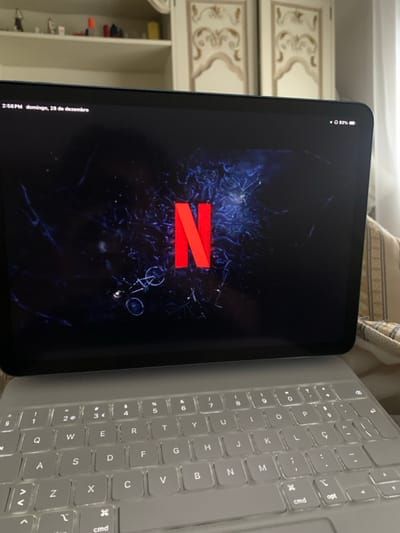 The image captures an electronic tablet, likely an iPad, positioned on a light grey keyboard case in an indoor setting within Macedo de Cavaleiros, Portugal. The tablet's screen displays the Netflix application's loading interface, featuring the prominent red "N" logo centered against a dark, abstract background with blue lighting effects and subtle, broken outlines resembling bicycles. The top left corner of the screen indicates "2:56 PM domingo, 28 de dezembro" (2:56 PM Sunday, December 28), while the top right corner shows a battery charge of 93%. The connected keyboard is a QWERTY layout with visible Portuguese character keys. In the background, a light-colored wooden cabinet with ornate, carved decorative panels is partially visible. To the left of the cabinet, a white shelf holds several small, indistinct objects. A striped, light-colored fabric element is visible to the immediate right of the tablet, and a white curtain or wall segment occupies the far right edge of the frame. No individuals are present in the scene.