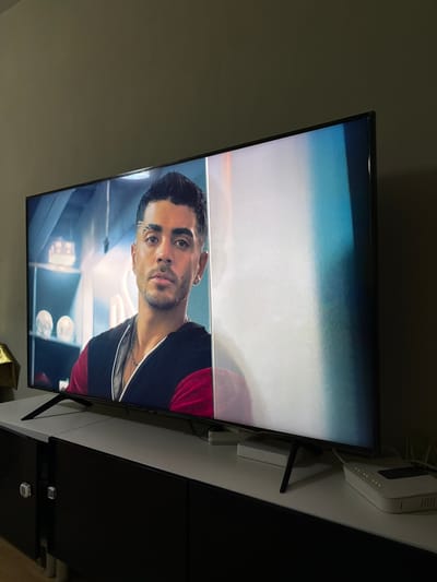 The image shows a large, black-framed television displaying a man's face in profile, set against a blurred background. The left side of the screen features the man, who has dark hair styled with a side part and a visible eyebrow slit. He is wearing a dark top with red accents and multiple necklaces. The right side of the screen is predominantly white with streaks of blue and pink, suggesting a visual distortion or a different scene entirely. The television is positioned on top of a modern, dark, two-door cabinet with silver handles. To the right of the television, a white modem-router with illuminated indicator lights and various cables is visible. The overall environment appears to be an indoor setting, likely a living room, with a neutral-colored wall in the background. The specific location of Getafe, Spain, is not depicted or suggested by any elements within the image.