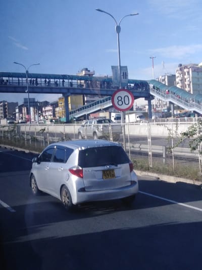 A silver hatchback is seen driving on a road alongside a metal fence. Beyond the fence, an elevated pedestrian walkway is visible, populated with people. A white and red circular sign displays the number 80. In the background, urban buildings stretch across the horizon under a clear blue sky. The car has license plate KCX 371P.