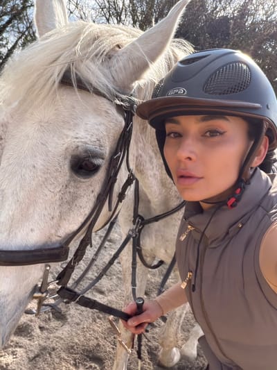 A person wearing a riding helmet and gray vest is seen in a close-up shot while on a white horse. The horse is wearing a bridle and bit. The background includes a blurry, leafless tree. The location is Grad Labin, Croatia.