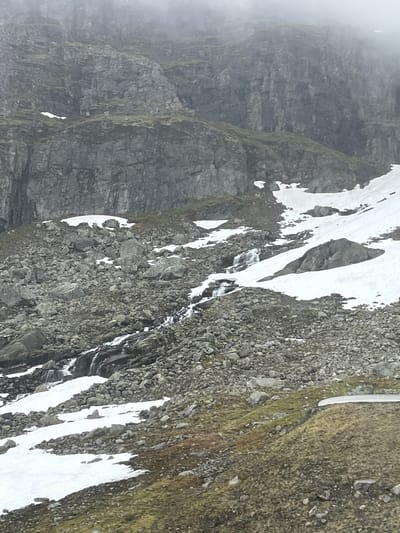 This image depicts a rugged, mountainous landscape with patches of snow scattered across steep, rocky slopes. The terrain is characterized by large, dark gray rock formations, some of which appear sheer and vertical, while others are covered in scree. Patches of low-lying green vegetation are visible on some of the less steep areas and clinging to the rock faces, suggesting a damp environment.
The snow appears to be melting, as evidenced by the streaky lines of water running down the rocks in several places, forming small rivulets. The overall atmosphere is cool and possibly damp, with a soft, diffused light suggesting an overcast or misty day. Fog or mist is visible in the upper left portion of the image, adding to the sense of elevation and remoteness. The scene is devoid of any human presence or man-made objects, focusing entirely on the natural geological features and the interplay of rock, snow, and water. The textures are rough and uneven, with the sharp edges of the rocks contrasting with the softer, more rounded forms of the snow and vegetation. The color palette is dominated by grays, whites, and muted greens, with the dark rocks forming the primary structural elements of the composition.