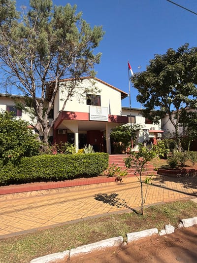 The image shows the exterior of a building with a sign that reads "POLICIA NACIONAL DIRECCIÓN GENERAL DE TALENTO HUMANO DIRECCIÓN DE GESTION DE PERSONAL". This suggests it is a police station or related administrative building. The building is painted cream and red, with a tiled walkway leading up to it. There are trees and bushes around the building, and the sky is clear and blue, indicating it is daytime with good weather. The sun casts shadows from the trees onto the walkway, suggesting it is mid-morning or afternoon. In the background, a flag is visible, partially obscured by trees. The overall scene depicts a government building in a well-maintained outdoor setting.
