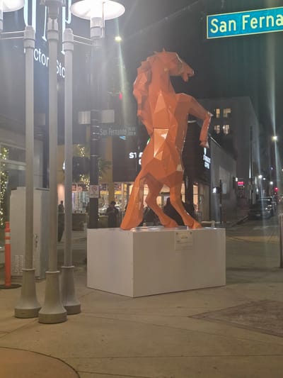 An orange, geometric sculpture of a horse stands on a white pedestal in front of a building at night, on the corner of San Fernando Boulevard and Palm Avenue in Burbank, California. The sculpture is lit by streetlights and the lights from the surrounding businesses. A street sign indicating "San Fernando BLVD 200 N" is visible above and to the right of the sculpture. A Starbucks Coffee store is located in the background to the right of the sculpture. Several people are visible in the background, some walking and others standing. The area is paved with concrete sidewalks and has streetlights illuminating the scene. The overall impression is of an urban streetscape at night, with the sculpture being the focal point.