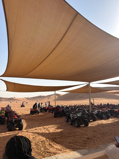 The photograph captures a scene in Al Madam, United Arab Emirates. A series of ATVs are parked on sand near an outdoor shelter made of tan fabric. People are standing near the vehicles. The environment appears to be a desert setting.