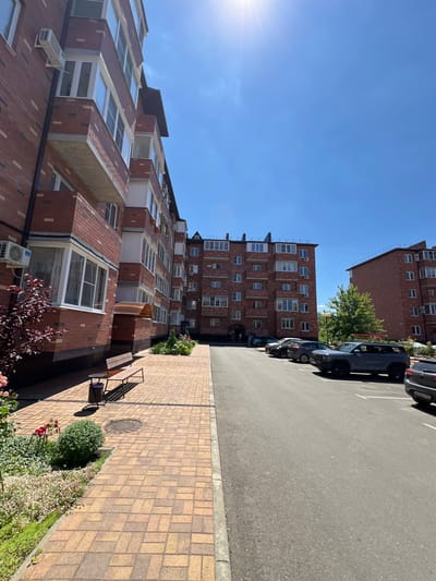 This is an exterior, eye-level, full shot of a residential area in Novaya Adygeya, Russia, on a clear, sunny day. The scene features a modern, multi-story apartment building constructed with red brick. The building has numerous windows and balconies. In front of the building, there is a paved walkway with a bench and a trash receptacle. Adjacent to the walkway is a small garden area with plants and flowers. The walkway leads to a paved road where several cars are parked, including a dark gray SUV in the foreground. Another apartment building is visible in the background. The sky is clear blue with the sun shining brightly. There are no discernible people actively engaged in interactions within the frame.