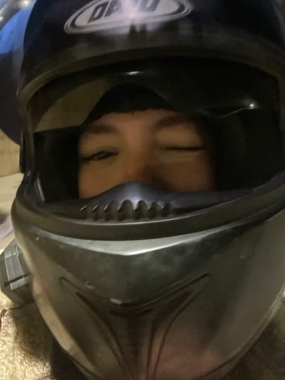 The image captures a close-up, frontal view of an individual wearing a dark full-face motorcycle helmet, located in Mechouar Fes Jdid, Morocco. The helmet, primarily black or dark grey, features "DAO" text branding on its top front. The chin bar includes a molded ventilation ridge. Only the individual's upper face is visible through the helmet's open visor: eyes, eyebrows, and the bridge of the nose. The individual's right eye (viewer's left) is open, while the left eye (viewer's right) is closed, suggesting a wink or similar expression. A dark fabric, possibly a balaclava or hair, is visible above the eyes. The background is dark and out of focus, with a faint, light-colored, textured element visible at the bottom left, and a blurred blue object in the upper left. The lighting is low.