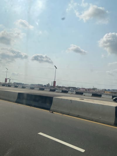 The image displays an elevated multi-lane asphalt road, presumably a flyover, under a partly cloudy sky within Ebute-Metta, Nigeria. The road surface is dark gray, featuring a prominent solid white lane marking in the lower-left foreground and a solid yellow line bordering a concrete barrier on the right. This primary barrier exhibits a black lower section and a light gray upper section. Further concrete barriers are visible along the road, some with alternating black and white segments. Tall streetlights, each topped with a flat, rectangular solar panel, are spaced along the bridge structure, two of which display red vertical banners. The background reveals a dense urban cityscape composed of various buildings, with a construction crane visible on the far left horizon. A small, dark-colored vehicle is partially visible on the far right, positioned behind the concrete barrier. No individuals are discernible, and no direct actions or interactions are taking place.