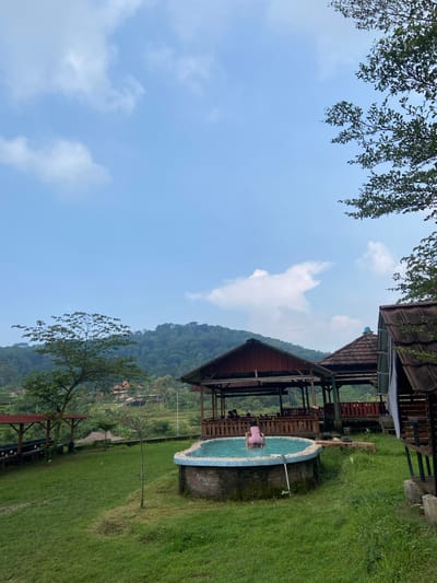 The image shows an outdoor scene near Penanggungan, Indonesia. The view includes a partially obscured mountain in the background and a blue and white tiled swimming pool. A wooden structure with a brown roof is adjacent to the pool. Lush green grass covers the ground. The sky is blue with scattered white clouds. Tree branches frame the top left corner. A person is partially submerged in the pool.