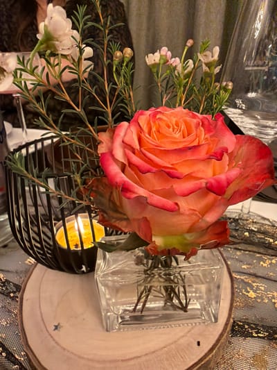 An eye-level shot showcases a peach-colored rose with red edges, accompanied by sprigs of tiny white flowers. The arrangement sits in a black wire basket, next to a small square glass vase on a wooden circle. A lit candle within the basket provides warm illumination. The background features a patterned fabric, adding depth and texture to the composition.