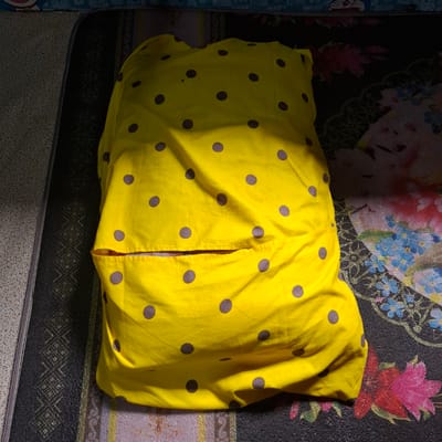 The image shows a yellow polka-dotted pillow placed on a patterned surface, likely a bed or mattress. The pillow is mostly covered by sunlight, creating strong shadows that obscure some of its details. The patterned surface beneath the pillow appears to be dark with floral motifs in shades of pink, blue, and red, and also has a black band with a geometric woven pattern along the left side. There is also a hint of a lighter, speckled pattern on the far left of the image. No people or actions are visible. This scene is set in an indoor environment, possibly a bedroom, in Reudeuep, Indonesia.