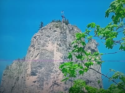 The image shows a rocky peak with a cross on top, against a bright blue sky. Green leaves from a tree in the foreground partially obscure the view of the peak. The rocky surface of the peak is textured and appears to be made of stone. A flag is visible on the cross. The overall scene is outdoors and appears to be a natural landscape. The location is identified as Oberviechtach, Germany.