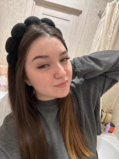 A young woman with brown hair, wearing a grey sweatshirt and a black headband, poses for a selfie in what appears to be a bathroom. Her left arm is bent behind her head. In the background, a cream-colored shower curtain is visible, along with bottles of toiletries on a shelf next to a bathtub. The overall lighting suggests an indoor, possibly domestic setting.