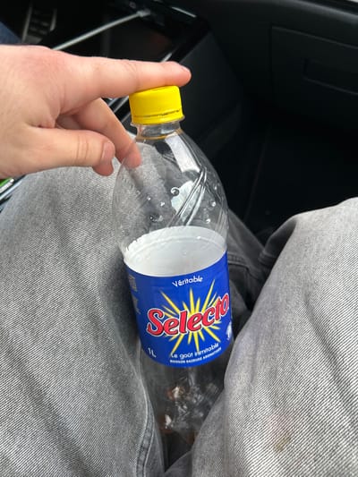 This image depicts the interior of a vehicle, likely situated in Hœrdt, France. A clear, 1-liter plastic bottle of "Selecto" carbonated beverage is centrally positioned. The bottle is largely empty, containing a small volume of dark liquid residue at the base, and is capped with a bright yellow screw lid. Its blue label displays "Véritable Selecto" in prominent red and yellow text, noting "1L" and "Le goût inimitable - BOISSON GAZEUSE AROMATISÉE". An adult's left hand is observed holding the upper neck of the bottle, just beneath the cap. The individual wears light grey denim trousers, which appear on either side of the bottle. A faint reddish-brown stain is visible on the right pant leg. The background consists of dark, molded plastic components, characteristic of a vehicle's interior dashboard or console.