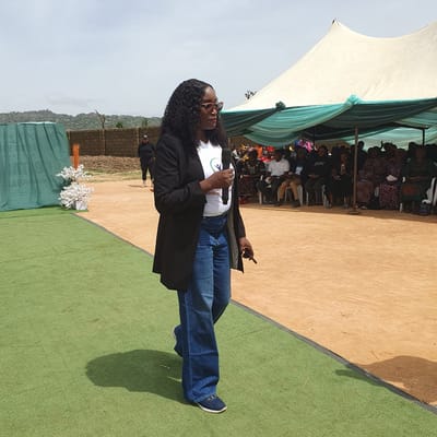 In Unguwar Saya, Nigeria, a woman stands on a green, artificial turf surface, holding a microphone in her right hand and a mobile phone in her left. She is wearing a white t-shirt with a green logo, a black blazer, and flared denim jeans, along with sunglasses and appears to be speaking. Behind her, a large tent with white and green fabric is set up, providing shade for a seated audience of men and women. The background shows a brick wall and a hilly, natural landscape under a cloudy sky. The overall scene suggests an outdoor event or gathering.