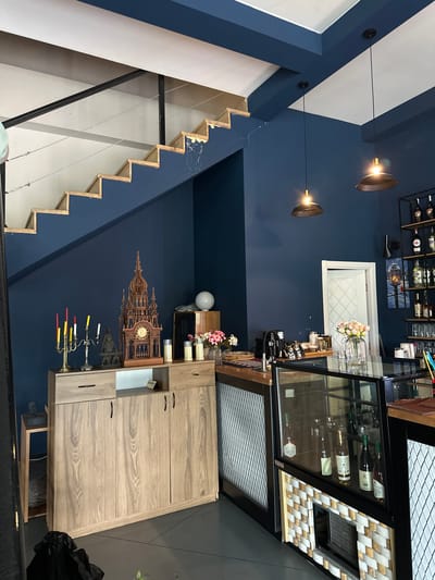 The image presents an interior view of what appears to be a business establishment, possibly a wine bar or boutique, situated in Marneuli, Georgia.
The decor features a modern aesthetic with dark blue walls contrasted by white architectural elements, most notably a staircase along the left side of the frame. Two pendant lights hang from the ceiling, illuminating the space.
On the right, a wooden cabinet with multiple drawers and cabinets stands against the wall. Above it, a display case with glass windows showcases wine bottles and glasses. Additional decorative items, such as flowers, candles, and an ornamental clock, are arranged on shelves and surfaces.
A visible rack of bottles mounted on the wall, reinforces the impression of a business serving alcohol, potentially wine. Overall, the scene depicts a well-maintained and thoughtfully decorated interior space in Marneuli, Georgia.
