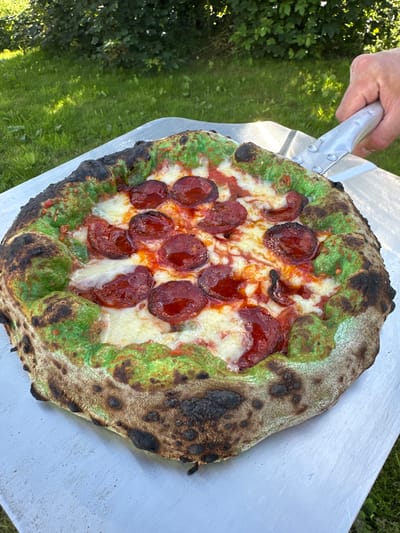 An overhead, close-up shot shows a pepperoni pizza on a pizza peel. The pizza has a bright green crust and is topped with pepperoni slices and melted white cheese. The pizza peel is silver and has a handle held by a person's hand which is visible on the top right of the image. The background consists of a grassy area with trees. The setting appears to be outdoors, in a backyard in Moortown, Ireland.