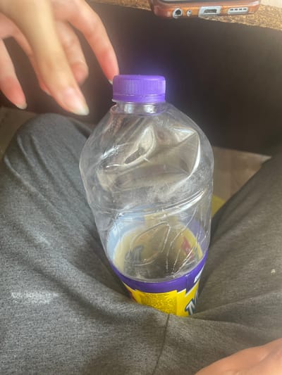 A person's hands are visible in the upper left corner, reaching towards a clear plastic bottle with a purple cap. The bottle is partially crushed and contains a yellow liquid. The bottle is resting on a person's lap, who is wearing grey sweatpants. The background is dark and out of focus, with a hint of a phone in the upper right corner. There is no indication of the location in the city of Besagash, Kazakhstan.