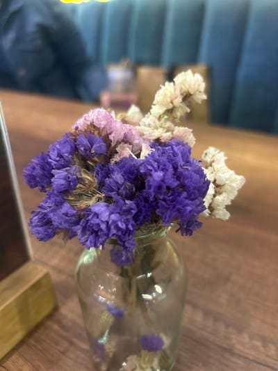 The image shows a small glass vase containing a bouquet of dried flowers. The bouquet features a mix of purple, pink, and white blooms. The vase sits on a wooden surface. The background is blurred but suggests an interior space. There is no evidence to confirm the location. It is impossible to determine if the location is in the city of La Aurora, Ecuador. There are no people, actions, or interactions visible in the photograph.
