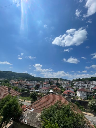 This media file presents a scenic view of a town nestled on a hillside under a bright, partly cloudy blue sky. The architecture is a mix of traditional tiled roofs and more modern, multi-story buildings, creating a sense of depth and history. Green trees and foliage are interspersed throughout the town, cascading down the slopes of the hill. The sun is shining, indicated by the bright illumination of the buildings and the presence of lens flare in the upper left portion of the image. The clouds are scattered and white, suggesting fair weather. The perspective is from a high vantage point, looking down and across the town, offering a panoramic vista. There are no people or specific activities depicted, making it a still-life landscape. The overall impression is one of a peaceful and picturesque setting, likely during the daytime.