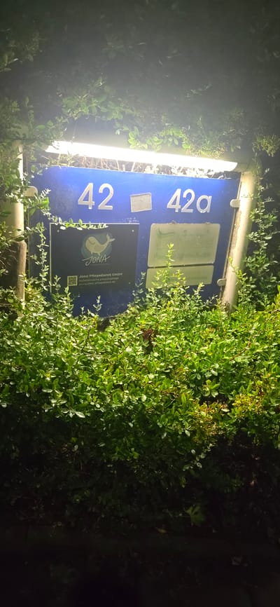 The image shows a blue sign with white numbers "42" and "42a" indicating a house or building number. The sign is mounted on a pole and partially obscured by green foliage and branches. Below the house numbers, there are two smaller signs. The left sign displays a logo of a whale and the text "JONA" and "Pflegedienst GmbH". Below this, there is a QR code and the website address "www.jona-pflegedienst.de". The right sign appears to be for a dental practice with the name "Wiedwild" and "Praxis für Zahnheilkunde". The scene is dimly lit, suggesting it is nighttime or dusk. The environment appears to be outdoors in an urban or suburban setting, with the foliage suggesting a garden or landscaping around the sign. The location is Seevetal, Germany.