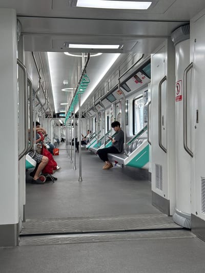 The image, initially rotated 90 degrees clockwise, depicts the brightly lit interior of a modern subway or train car, likely in Tiaodeng, China, as indicated by the context. The train car features a clean aesthetic dominated by white and light grey surfaces, accented with a distinct mint green on the seating and some fixtures.
The scene shows several passengers engaged in travel. In the mid-ground, a young person wearing a grey top and dark pants is seated, looking downwards, possibly at a phone or book. Further into the background, other passengers are visible, some seated and some standing, though their details are less clear. In the foreground, partially obscured, a person is seated with their legs extended, appearing to rest or sleep, with a red bag or trolley next to them. The overall atmosphere appears calm and unhurried.
The interior is well-appointed with numerous stainless steel handrails running along the ceiling and vertically between seating areas, along with mint green hanging straps for standing passengers. Long, linear LED light strips illuminate the ceiling and upper walls, contributing to the bright environment. The floor is light grey, with a textured, possibly non-slip, surface, and a section near a door features a diamond-plate metallic pattern. Overhead, white panels integrate air vents.
A notable detail is a red sign with white Chinese characters visible above a seat, which reads "禁止倚靠车门" (Jìnzhǐ yǐkào chēmén), meaning "No leaning on the car door." This text confirms the location within a Chinese-speaking region and reinforces its identity as a public transport vehicle. The car design, including the specific color scheme and modern fixtures, is characteristic of contemporary subway systems.