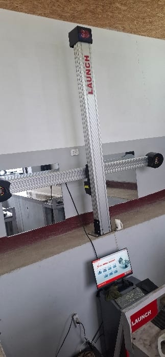 An automotive wheel alignment system by Launch is positioned within a garage. The system features a horizontal bar with "LAUNCH" printed on it, supported by two vertical poles. A computer console with a display screen is located to the right of the equipment. The console's display presents various vehicle-related data and graphics. A power outlet is visible on the wall.