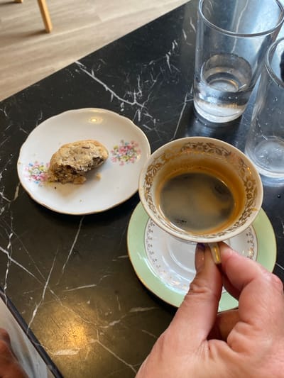An overhead shot depicts a black marble-patterned table in La Tour-d'Aigues, France. On the table, a partially consumed, light-colored cookie with dark inclusions rests on a small white plate with a floral pattern and gold rim. To the right, two clear glass tumblers are present; one contains a small amount of clear liquid, while the other is mostly empty. A white ceramic demitasse cup with gold detailing and a gold handle, filled with dark coffee topped with crema, is held by a hand with light-colored fingernails, lifting it from its light green and gold-rimmed saucer. A light-colored leg or garment is visible in the lower left quadrant of the frame, indicating a person is seated at the table. A light-toned wooden floor or surface is visible in the blurred background.