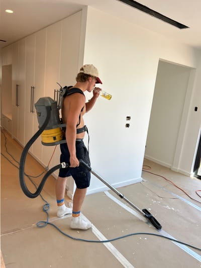 A shirtless man, wearing a cap, shorts, and socks, drinks from a beer bottle while operating a backpack vacuum cleaner. He is angled as if reclining, vacuum wand in hand. The room is under construction, with paint lines on the floor, bare walls, and newly installed cabinets. The overall scene suggests a casual or humorous approach to a cleaning task in a modern, unfinished space.