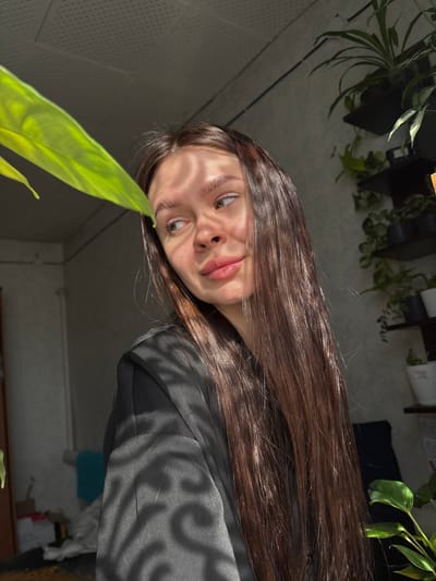 A young woman with long brown hair is the primary subject of the image. She is wearing a black jacket. The background shows a room with plants on shelves. A green leaf of a plant is in the foreground. The scene appears to be indoors, with sunlight casting shadows. There is no evidence to confirm a location in the city of Dubrova, Russia.
