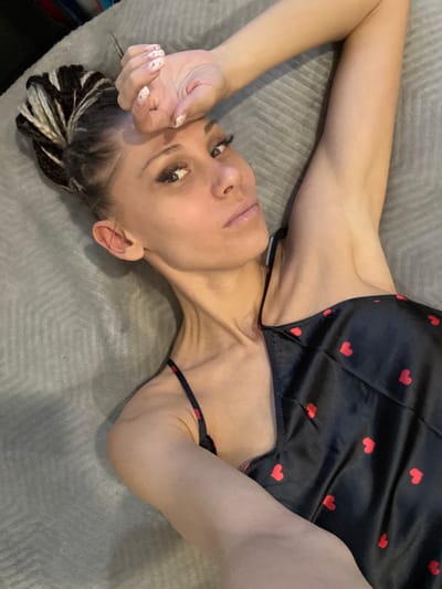 A woman lies on a grey surface, possibly a bed or sofa. She is wearing a black top with red heart patterns. Her hair is styled in braids with some white strands. Her left hand is near her forehead, displaying painted nails. The environment lacks context to determine a location in Yaroslavl', Russia.