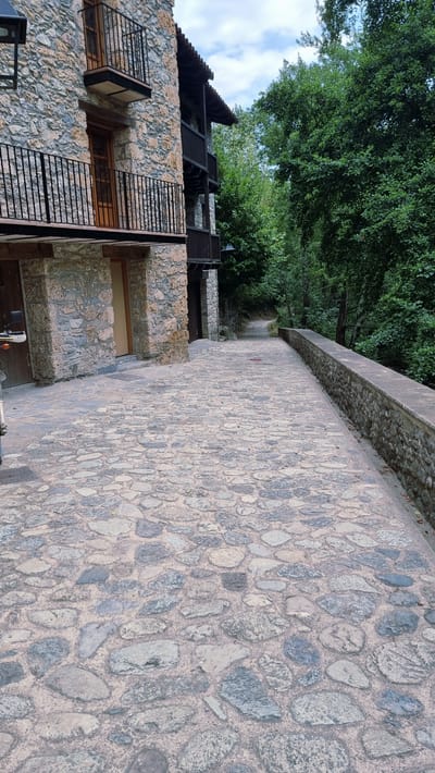 An eye-level, medium shot captures a cobblestone pathway leading away from a stone building in Camprodon, Spain. The pathway is paved with irregular grey and pink stones of varying sizes, with a concrete wall on the right side. The stone building, to the left, features two balconies with black railings and a wooden door. Lush green trees line the right side of the pathway, creating a natural backdrop. The sky above is partly cloudy. No people or animals are visible.
