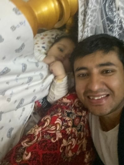 A man and baby are lying on a bed in Galaosiyo, Uzbekistan. The man is smiling at the camera. The baby is wrapped in a blanket labeled "Baby". A portion of a golden cylindrical object is visible on the left. The bed has a red and black floral pattern.