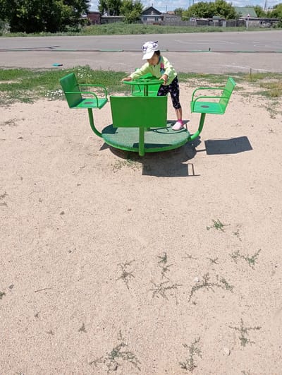 In a sandy outdoor area in the city of Zhaspáy, Kazakhstan, a young girl wearing a light green jacket, dark patterned leggings, and pink sandals stands on a green, circular merry-go-round. She is wearing a white cap with cartoon designs. The girl is holding onto the central steering mechanism of the merry-go-round. The merry-go-round has two green seats, one to her left and one to her right. The ground is covered in light-colored sand with scattered small green weeds. In the background, there is a paved area with white markings, a line of green grass, trees, and several buildings, suggesting a residential or community area. The sky is cloudy.