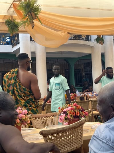 The image displays an outdoor social gathering located in Kumasi, Ghana. Overhead, light orange fabric drapes a canopy structure, accented with suspended artificial green foliage in bamboo-like holders. In the background, a multi-story building features white architectural pillars, green walls, and dark-framed windows, with an upper-level balcony showcasing black and gold ornamental railings.
In the immediate foreground, partial views of two older men, both with dark skin and bald heads, are visible, seated at tables. The man on the left is seen from his back and left shoulder. The man on the right, wearing a light-colored shirt, is viewed from the back of his head and right shoulder, facing left with his mouth slightly ajar.
The middle ground features three standing men. On the left, a man with a fade haircut and a short beard wears dark sunglasses and a traditional green, gold, and red patterned kente cloth wrapped around his torso, leaving his right shoulder bare. He faces away from the camera. Center-frame, a man with short dark hair and dark skin wears a mint green short-sleeved top with a teal accented chest pocket, looking directly at the camera with a neutral expression. To his right, a third man, similarly dressed in a mint green top, stands with his right hand near his head.
The gathering is furnished with woven wicker-style chairs arranged around tables draped with light-colored tablecloths. Each table is set with clear glass plates and features potted artificial flower arrangements primarily in pink, orange, and red tones. Additional guests are visible seated at tables in the blurred background.