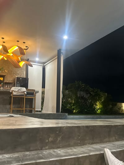 The scene depicts a nighttime view of an outdoor or semi-outdoor area within a property located in Tumbak Bayuh, Indonesia. The upper left portion shows a white ceiling with multiple recessed spotlights and five distinct, curved, wooden pendant light fixtures emitting a warm, orange-yellow glow. Below these, a textured, light-brown wall serves as a backdrop to a dark-colored bar counter. On the counter, a small, black refrigerator is visible, along with several glasses. A wooden bar stool, with a white t-shirt draped over its back, stands in front of the counter. To the right of the bar, two tall, light-colored roll-down screens or blinds are partially lowered.
The right side of the image features a completely dark night sky above a lush, green arrangement of tropical vegetation, including various plants and small trees, which are illuminated from below by multiple yellow-green spotlights. Behind the vegetation, a light-colored wall with square, decorative cut-outs is partially visible. In the middle ground, a dark, still body of water, likely a swimming pool, extends across the width of the frame between the covered area and the garden.
The foreground is dominated by broad, rough-textured concrete steps or platforms, indicating a multi-level floor plan. A white cloth or towel lies on one of these concrete surfaces near the bar area. In the bottom right corner, the toe of a white athletic shoe is partially visible, indicating the presence of an individual outside the main focal points of the scene. No active interactions or specific actions are depicted.