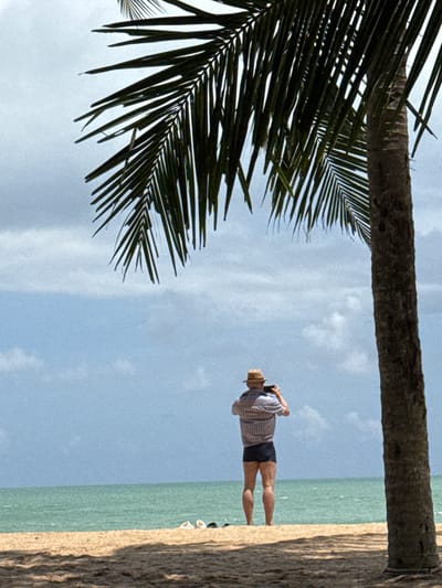 A man wearing a straw hat and a plaid shirt stands on a sandy beach in Ban Khuk Khak, Thailand, holding a black camera to his face. He is facing away from the viewer, towards the ocean. The man is wearing dark shorts. A palm tree looms to the right of the frame, its fronds extending over the scene. The ocean is a calm, turquoise color, meeting a hazy blue sky with scattered white clouds. Two pairs of flip-flops are visible on the sand in the distance. The sand itself has a rough texture, with shadows cast from the palm tree. The overall scene suggests a relaxed, tropical atmosphere.
