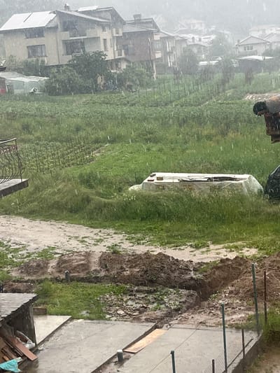 The image depicts an outdoor residential and agricultural scene in Elhovets, Bulgaria, under conditions of heavy rainfall. Numerous streaks of rain are visible across the entire frame, impacting visibility and rendering all surfaces wet.

In the background, a cluster of multi-story houses with pitched roofs is situated on a gentle slope. At least one house on the left-middle side of the background features multiple solar panels on its roof. Other houses further back are partially obscured by the rain and distance.

The mid-ground is dominated by extensive green fields or gardens, organized into discernible rows of vegetation, some supported by stakes, indicative of cultivation. To the far left, a small, translucent greenhouse structure is partially visible. In the central-right mid-ground, a long, rectangular object covered by a translucent white tarp rests on a low base amidst tall grass. A dark, weathered, possibly rusty structure is partially visible on the far right edge of the frame.

The foreground features a wet concrete slab or patio. To the left, a small, dark, shed-like structure is present, with wooden planks and other materials stacked nearby. Adjacent to the concrete, significant mounds of dark, wet earth are visible, indicating recent excavation, including a distinct trench. Puddles and muddy areas are widespread across the disturbed ground. A metal fence with green posts runs along the lower edge of the foreground. A decorative metal balcony railing extends into the frame from the far left, suggesting the image was captured from an elevated vantage point.

No individuals or animals are present in the scene. The overall lighting is diffuse, consistent with an overcast and rainy day.