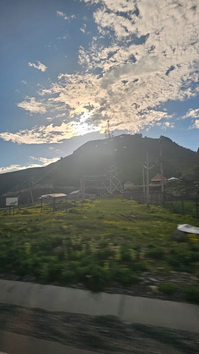 This is a photograph of a mountainous landscape with a bright sky filled with clouds, likely taken during the daytime. The image is rotated 90 degrees counter-clockwise.

In the foreground and midground, there is a steep, grassy hillside dotted with small structures, possibly houses or sheds, and a significant amount of electrical infrastructure, including power lines and tall transmission towers. The grass on the hillside appears lush and green, with some hints of yellow wildflowers. To the right of the hillside, a paved road or pathway runs alongside it. The sky is a clear blue with large, fluffy white clouds that are illuminated by sunlight, creating a bright and somewhat hazy effect, especially near the horizon where the sun is likely positioned.

The presence of power lines and transmission towers suggests this is an area where electricity is being transported, possibly a rural or mountainous region that requires such infrastructure. The small buildings on the hillside hint at human habitation or activity within this natural setting. The overall mood of the image is peaceful and serene, evoking a sense of natural beauty combined with human-made structures. There is no visible text in the image.