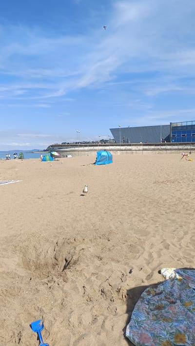 This is a photograph of a beach scene. The foreground is dominated by sandy ground, with a blue toy shovel visible in the bottom left corner. A seagull stands in the middle ground, looking towards the camera. Further back, there are several beach tents, one of which is blue and open. A patterned beach towel is draped on the sand in the bottom right. In the background, there is a wall with lampposts and a large building with a modern, angular design. Beyond the building, the sea is visible under a bright blue sky with scattered clouds. There are a few people visible on the beach near the water and along the wall. There is also a bird in flight high in the sky. The location is not Greci, Italy, but appears to be a seaside location with a developed promenade.