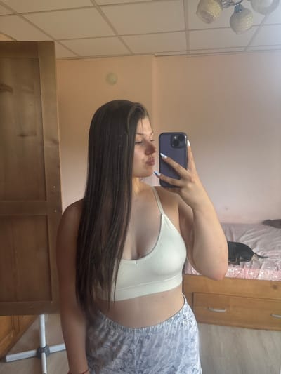 The media file shows a young woman taking a selfie in a mirrored room. She is wearing a white ribbed bralette and grey patterned shorts. Her long, dark hair is visible, as is part of her face and arm. In the reflection of the mirror, a dark-colored dog is visible on a pink blanket on a bed. A wooden wardrobe and a part of a dresser are also visible in the background. The room appears to be a bedroom or dressing room. There is no visible text in the media file. The lighting suggests it is daytime.