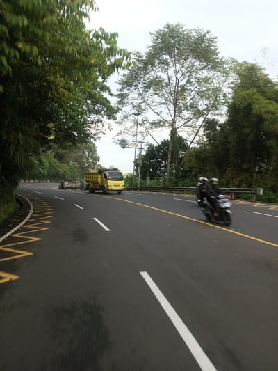 On a winding road lined with trees, possibly in Taunggyi, Myanmar, a yellow dump truck travels in the opposite direction. Two people on a motorcycle are heading towards the viewer. The road has white lane markings and a yellow line. A row of crash barriers is on the right-hand side of the road. The sky is overcast.