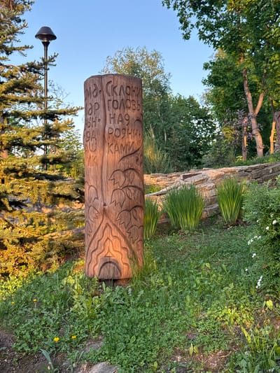 The image presents a horizontal, eye-level shot of a carved wooden log situated in a natural, outdoor setting. The log, cylindrical in shape, features carvings of leaves or foliage and inscription in the Russian language. The location is Krasny Klyuch, Russia.
The environment consists of lush greenery including various grasses and small plants. A rock wall or formation is visible in the background, and scattered tufts of elongated grass plants grow. Trees and a partly obscured lamp post are also visible. The sky is a clear blue gradient.