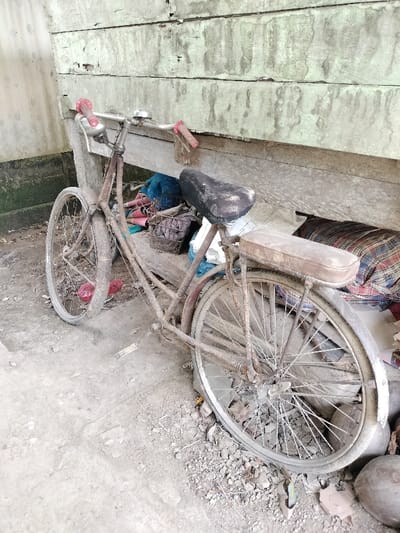 A rusty bicycle leans against a weathered wooden wall. The bike, showing its age, has flat tires, a worn seat, and peeling red grips. Assorted items are piled behind it, including bags and a woven basket. The ground around the bicycle is covered in dirt, debris, and a few coconuts. The wall behind shows signs of decay and peeling paint, hinting at neglect and the passage of time.