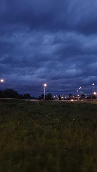 The image captures a scene in Obukhovo, Russia, at dusk or night. A field of dark green grass occupies the right side of the frame. A row of streetlights extends into the distance, illuminating the road running adjacent to the field. Beyond the streetlights, glimpses of buildings are visible. The sky is filled with dark, heavy clouds, suggesting an approaching storm. The overall impression is one of a quiet, overcast evening in a suburban or semi-rural area.