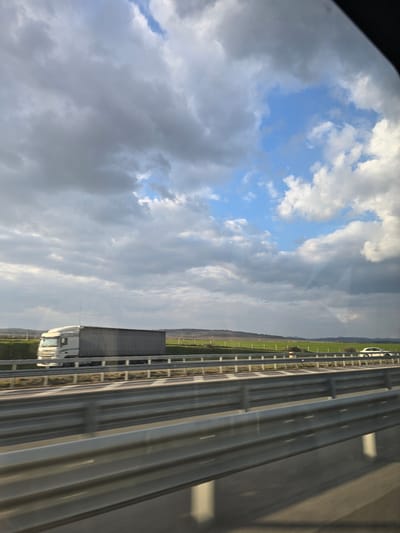 The scene is located in the vicinity of Vinarovo, Bulgaria, depicting a multi-lane highway under a dynamic sky. The upper portion of the image is dominated by a mix of heavy, dark grey storm clouds and patches of clear blue sky with white cumulus formations, through which visible sun rays descend onto the distant landscape. In the mid-ground, a highway stretches with multiple lanes, featuring grey metal guardrails running parallel to the direction of travel in the foreground and along a median. Shadows from these guardrails are cast onto the asphalt. A white semi-truck with a grey curtain-sided trailer is traveling on the far lane in the same direction as the viewpoint. Further in the distance, a white passenger car is visible on the opposing side of the highway. Beyond the highway and its immediate shoulder, the environment comprises expansive green fields extending towards rolling hills and a distant tree line under the varied sky conditions.