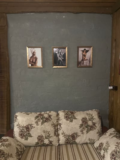 The image shows a wall with three framed photographs. The photographs depict nude women in vintage-style poses. The wall is painted a dark gray and appears to be textured, possibly brick. Above the wall is a wooden beam. Below the wall is a sofa with patterned cushions. The room appears to be dimly lit. The location is described as being in the city of Lapės, Lithuania, though this cannot be confirmed from the image alone.
