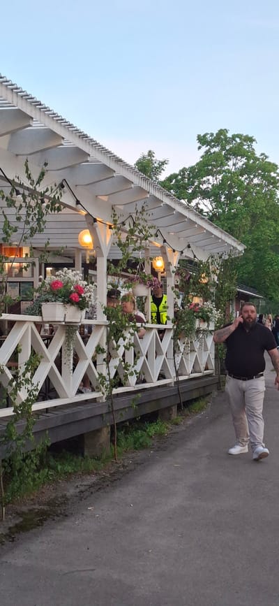 The image depicts an outdoor seating area of a restaurant or cafe, likely in the city of Salpakangas, Finland. The establishment features a white wooden gazebo with decorative latticework and hanging plants, illuminated by string lights. Several people are seated at tables under the gazebo, appearing to be patrons. A man in a black polo shirt and light-colored pants is walking on a paved path in the foreground, talking on his mobile phone. He has tattoos visible on his arm. Behind the gazebo, a red building and lush green trees are visible, suggesting a natural or park-like setting. The sky is clear and blue. The overall atmosphere appears casual and relaxed.