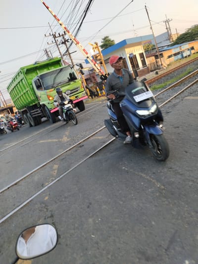 This image depicts a street scene in Bringinbendo, Indonesia, near a railway crossing. The time appears to be late afternoon or early evening, indicated by the warm, low-angled sunlight casting long shadows. The sky is clear and bright.
The main subjects are several vehicles and people. Dominating the left side of the frame is a large, bright green dump truck. In front of the truck, and to its right, are several motorcycles. A man wearing a red cap and a dark shirt rides a blue scooter in the foreground, positioned between the viewer and the truck. Another man on a motorcycle is visible between the truck and the blue scooter. Further back, more motorcycles are seen, some with passengers. A railroad crossing gate, painted with red and white stripes, is raised, allowing traffic to pass over the tracks. In the background, there's a small building with blue and white accents, possibly a shop or station, and overhead electrical wires are prominent.
The activity is traffic moving through the railway crossing. The presence of multiple motorcycles suggests a busy road. The overall mood is ordinary, depicting daily life and transportation. The man on the blue scooter seems to be looking to his left, perhaps anticipating the train or observing the traffic.
Visible text includes a license plate on the blue scooter which reads "B 6933 KBO". The green truck has "HINO" written on its front grille. The building in the background has the number "39" displayed. The weather appears to be clear and sunny. The ground is asphalt, marked with yellow lines, and the railway tracks are clearly visible. The reflection in a motorcycle's side mirror on the bottom left shows a person wearing a white collared shirt.